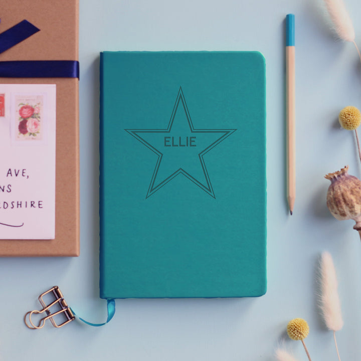 A flat lay image of a turquoise vegan leather notebook resting on a pale aqua table. The front of the notebook is engraved with a large 5 point star and is personalised with the name Ellie. Surrounding the book are various stationary items and dried flowers.
