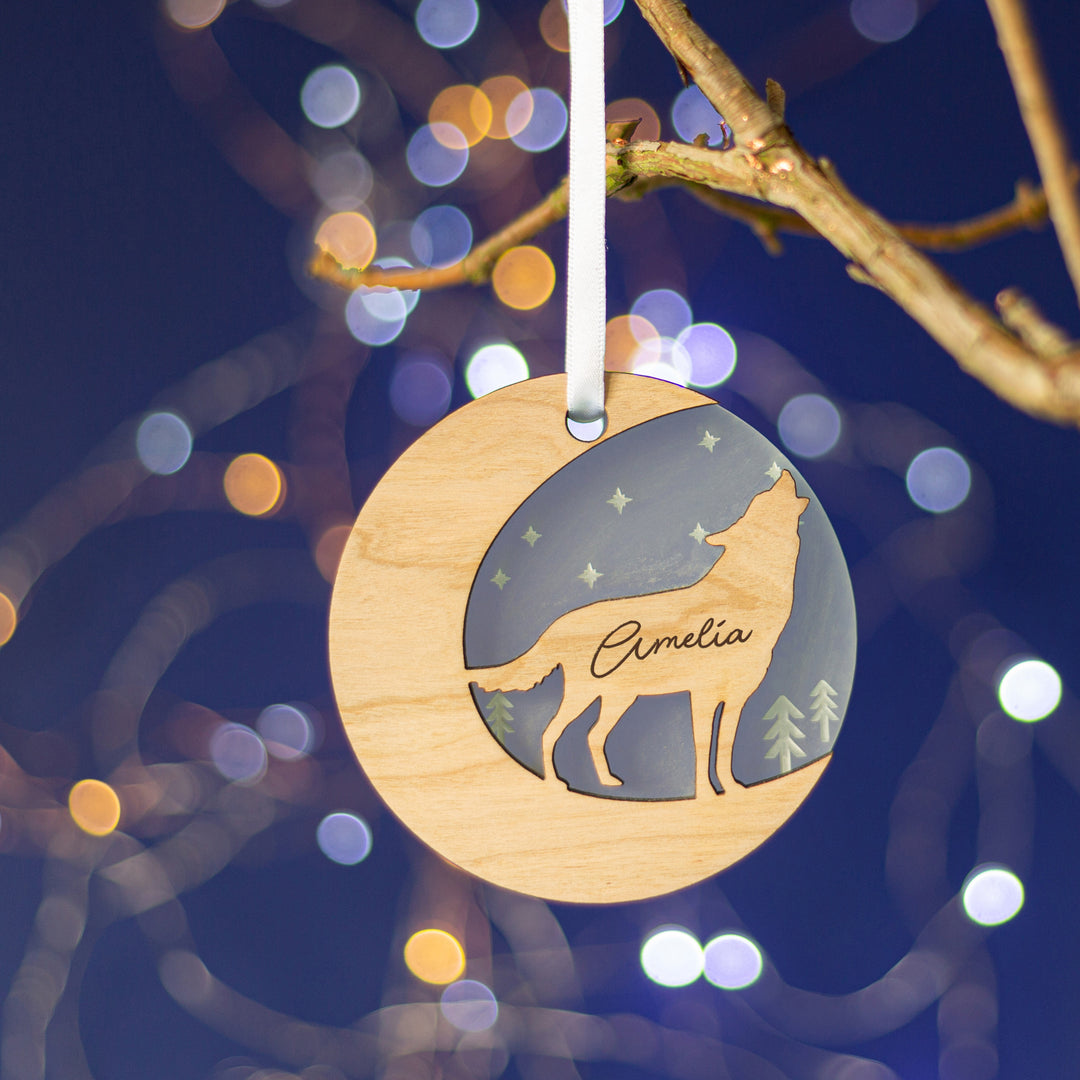 A wooden Christmas ornament shaped like a crescent moon hangs from a branch against a dark blue background with bokeh lights. The ornament features a cutout silhouette of a wolf howling at the moon and is engraved with the name "Xander."