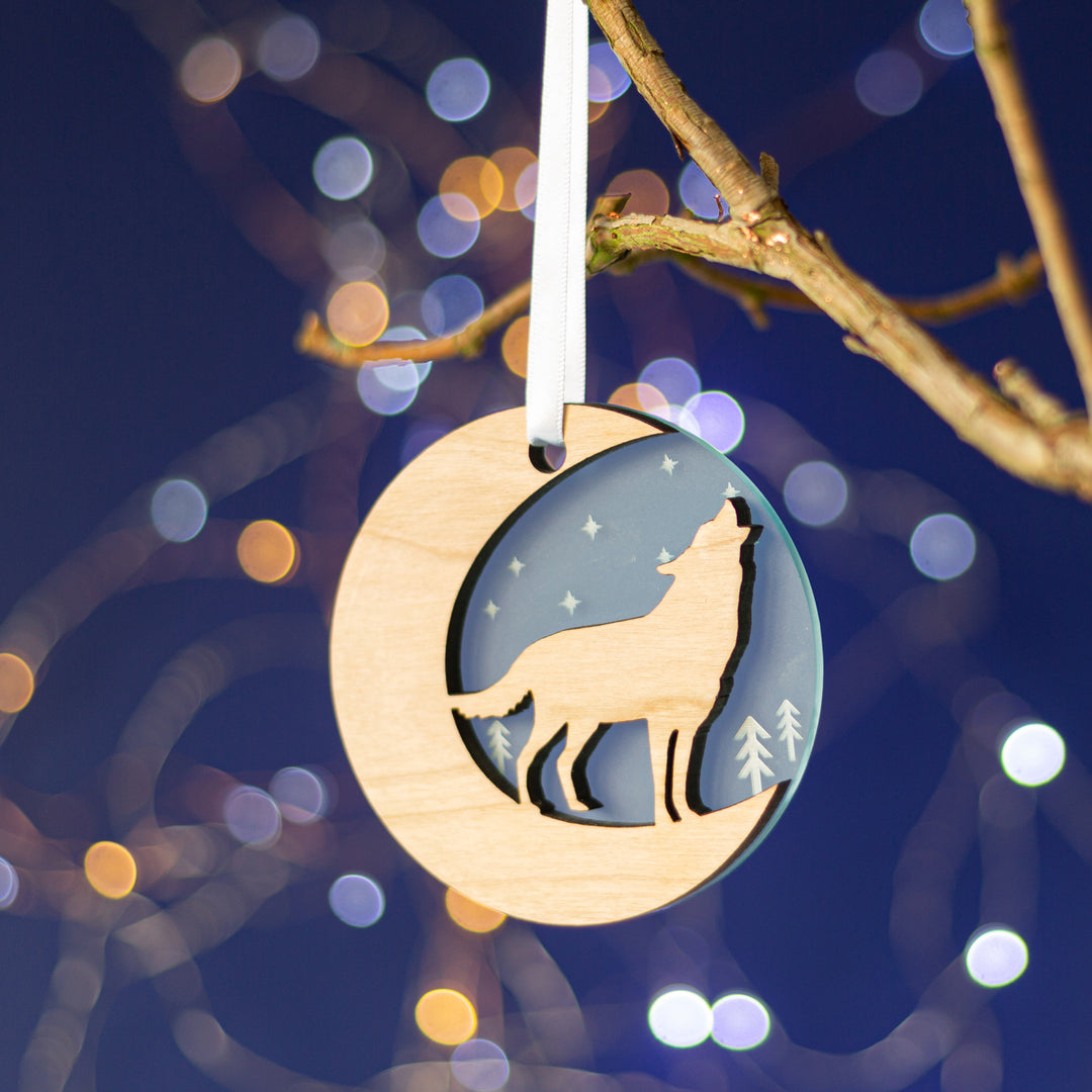  A round wooden Christmas ornament hangs from a branch against a blue background with bokeh lights. The ornament features a cutout silhouette of a wolf howling at the moon.