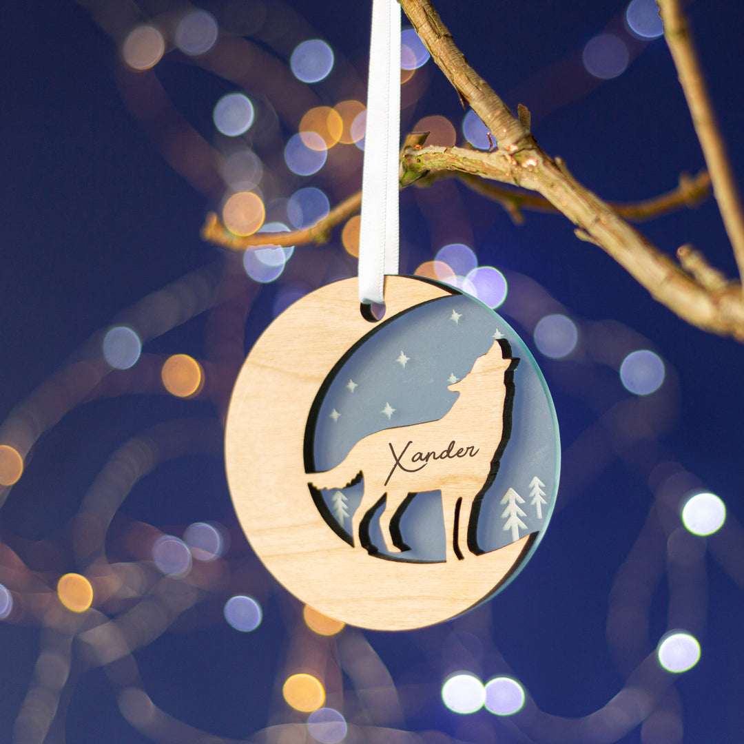 A light blue wooden Christmas ornament featuring a howling wolf silhouette against a starry night sky, personalized with the name "Xander," hanging from a tree branch with twinkling lights in the background.
