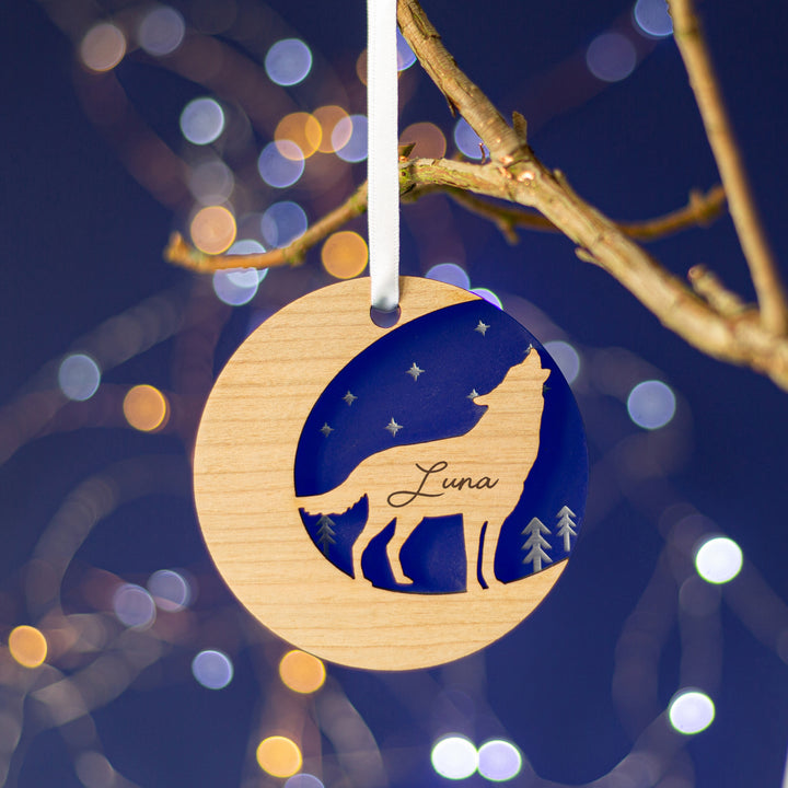 A round wooden Christmas ornament hangs from a branch against a dark blue background with bokeh lights. The ornament features a cutout silhouette of a wolf howling at the moon and is engraved with the name "Luna."