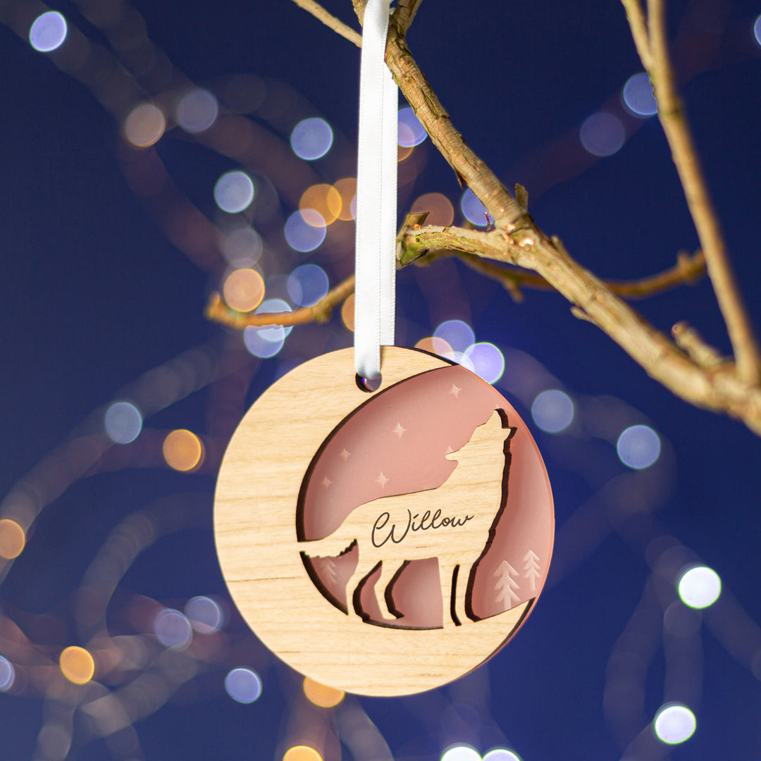 A wooden Christmas ornament featuring a howling wolf silhouette against a starry night sky, personalized with the name "Willow," hanging from a tree branch with twinkling lights in the background.