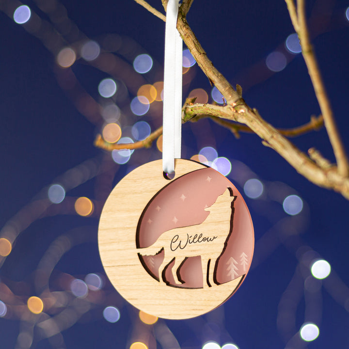 A wooden Christmas ornament featuring a howling wolf silhouette against a starry night sky, personalized with the name "Willow," hanging from a tree branch with twinkling lights in the background.