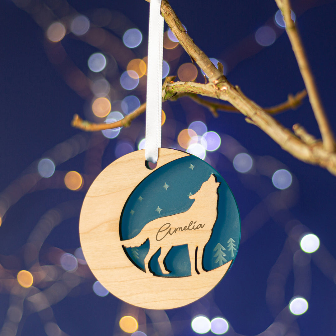 A teal wooden Christmas ornament shaped like a crescent moon hangs from a branch against a dark blue background with bokeh lights. The ornament features a cutout silhouette of a wolf howling at the moon and is engraved with the name "Amelia."