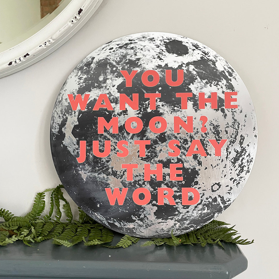 A round moon-themed wall plaque with the quote: "YOU WANT THE MOON? JUST SAY THE WORD" in bold red letters, displayed on a mantelpiece with green ferns.