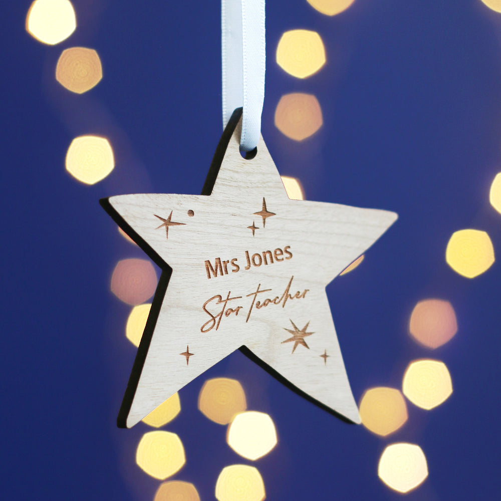 A wooden star-shaped Christmas ornament hangs against a dark blue background with bokeh lights. The star is engraved with the text "Mrs. Jones" and "Star Teacher" and has small star decorations around it.