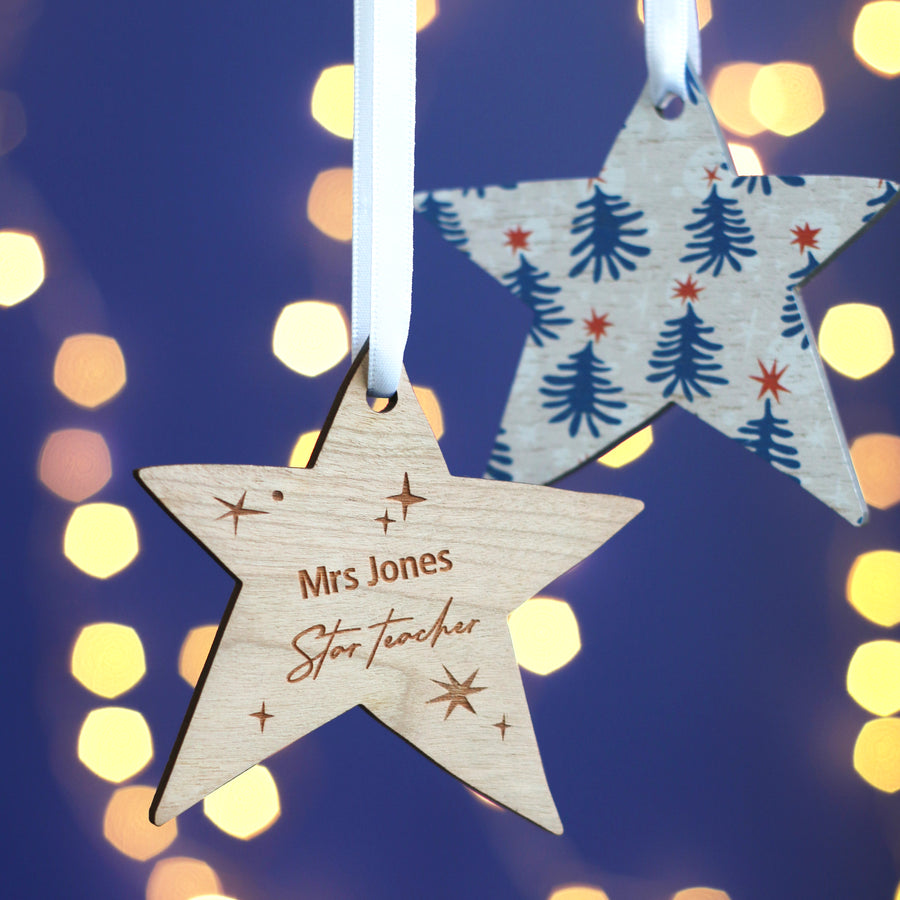 Two wooden star-shaped Christmas ornaments hang from a branch against a dark blue background with bokeh lights. One star is decorated with a pattern of blue trees and red stars. The other star is engraved with the text "Mrs. Jones" and "Star Teacher" and has small star decorations around it.