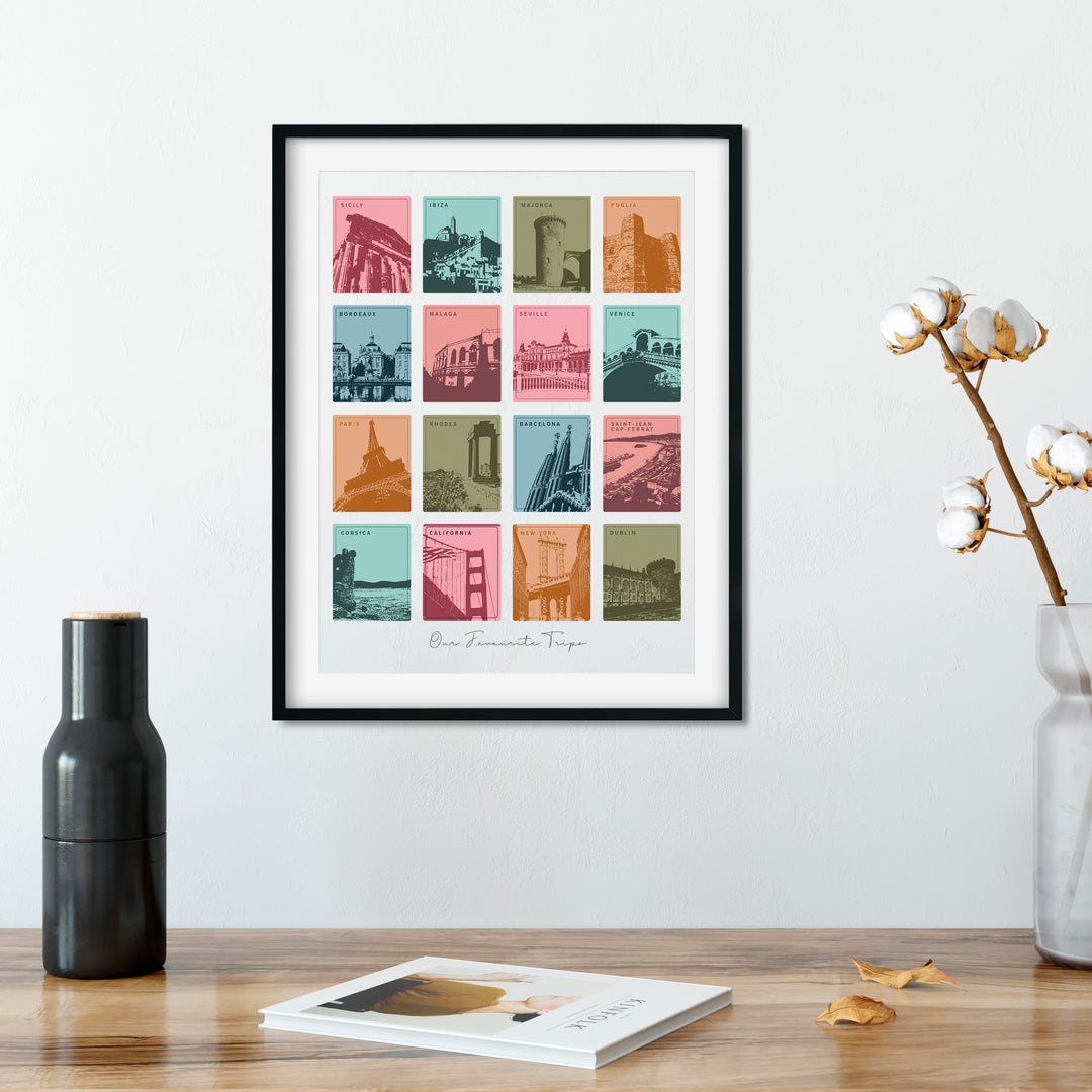 A framed print with 12 colorful squares, each depicting a different city landmark, hangs on a wall above wooden table. A black vase, a cotton branch in a clear vase, and a book are also on the table. The print is personalized with "Our Favourite Trips" and "KINFOLK" is visible in the book.