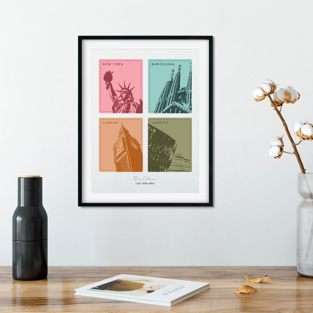 A framed print with four colorful squares, each depicting a different city landmark, hangs on a wall above wooden table. A black vase, a cotton branch in a clear vase, and a book are also on the table. The cities are New York, Barcelona, London, and Cardiff. The print is personalized with "Love from Annie."