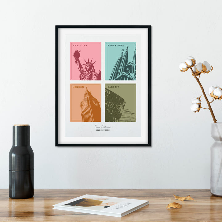 A framed print with four colorful squares, each depicting a different city landmark, hangs on a wall above wooden table. A black vase, a cotton branch in a clear vase, and a book are also on the table. The cities are New York, Barcelona, London, and Cardiff. The print is personalized with "Love from Annie."