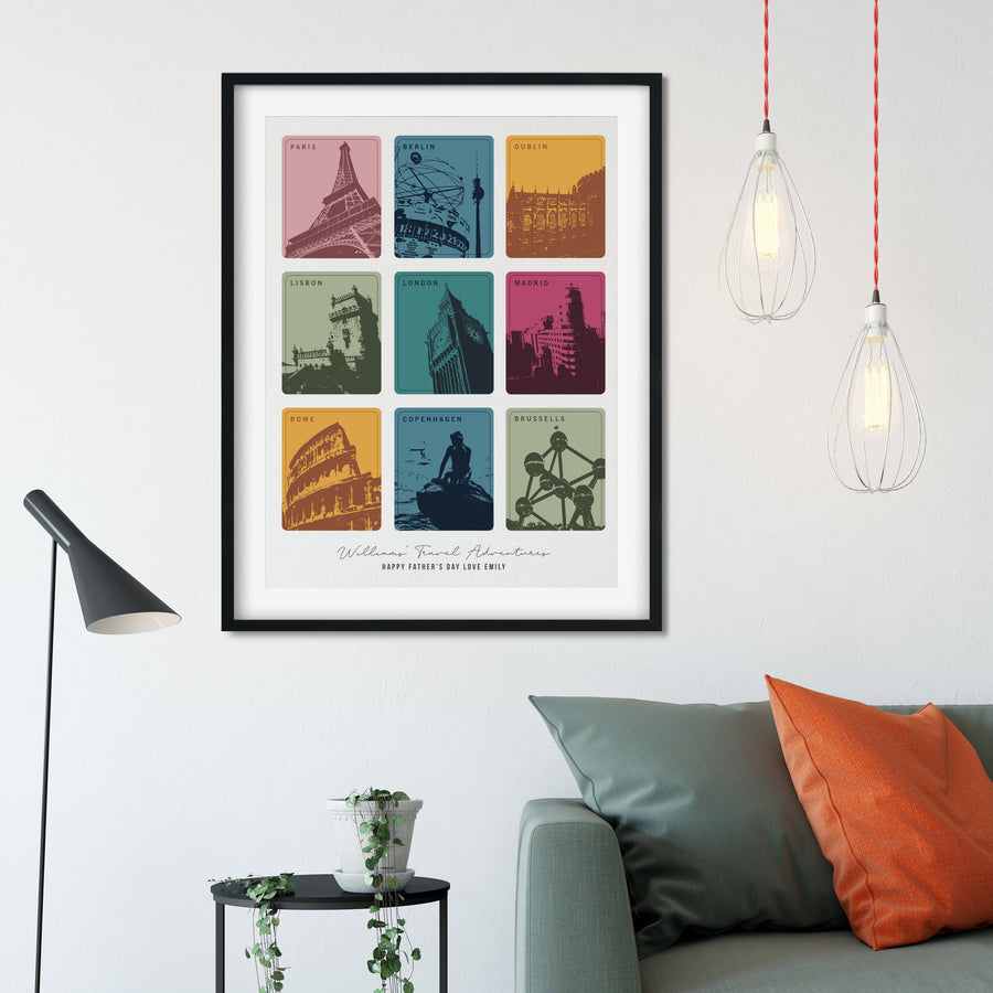 A framed print with nine colorful squares, each depicting a different European city landmark, hangs above a gray couch with orange pillows. The print is personalized with "William Travel Adventures" and "Happy Father's Day Love Emily." Two pendant lights hang above the couch.
