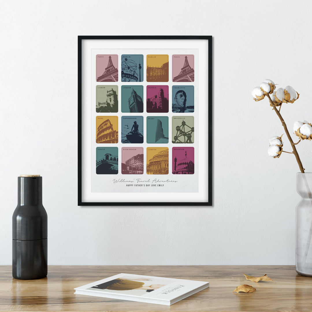  A framed print with 12 colorful squares, each depicting a different city landmark, hangs on a wall above wooden table. A black vase, a cotton branch in a clear vase, and a book are also on the table.
