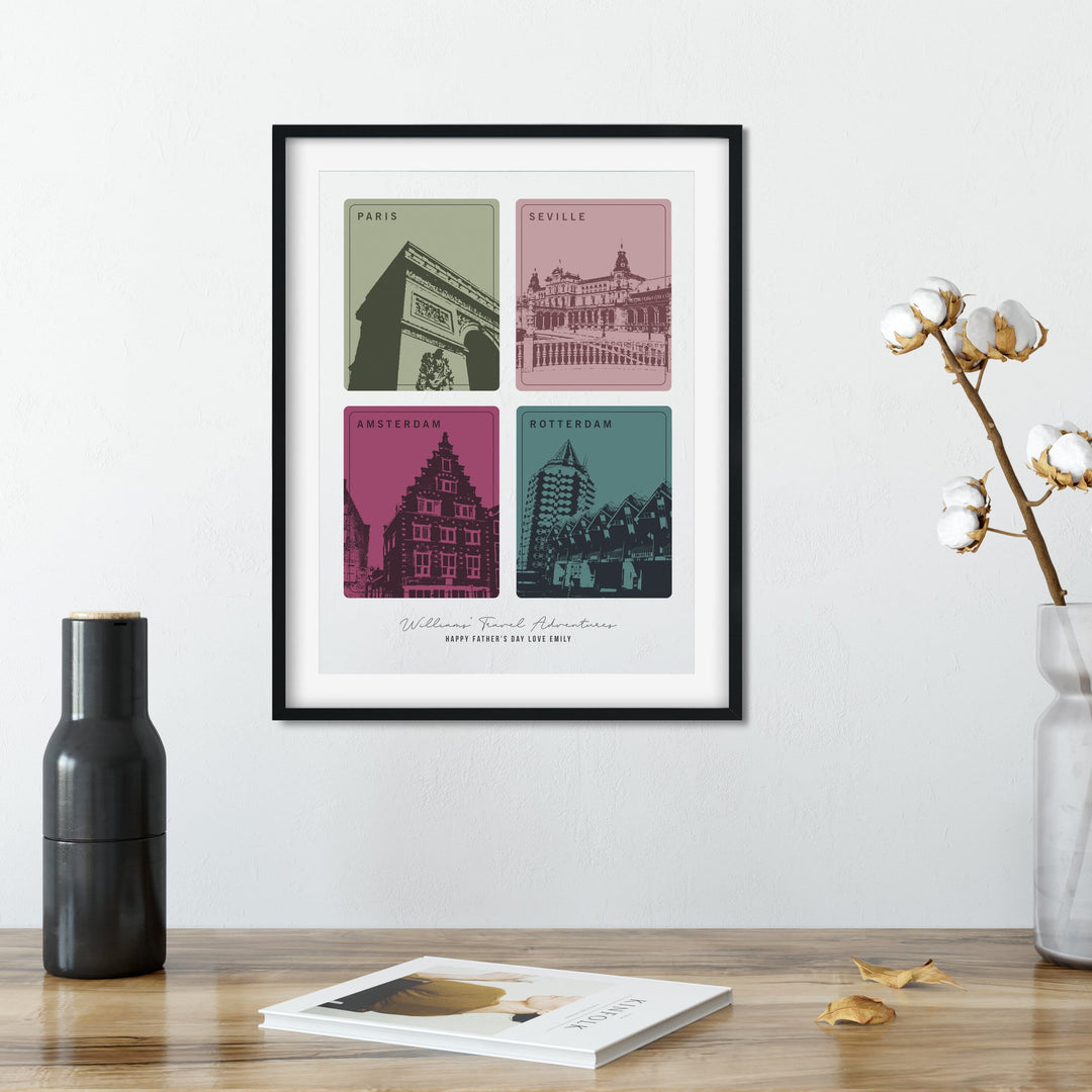 A framed print with four colorful squares, each depicting a different European city landmark, sits on a wooden table. A black vase, a cotton branch in a clear vase, and a book are also on the table. The cities are Paris, Seville, Amsterdam, and Rotterdam. The print is personalized with "Williams Travel Adventures" and "Happy Father's Day Love Emily."