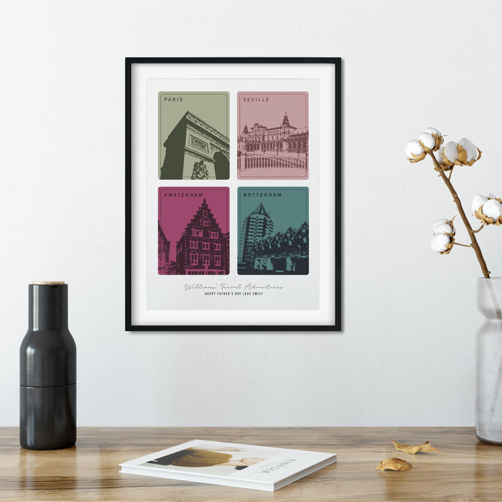 A framed print with four colorful squares, each depicting a different European city landmark, sits on a wooden table. A black vase, a cotton branch in a clear vase, and a book are also on the table. The cities are Paris, Seville, Amsterdam, and Rotterdam. The print is personalized with "Williams Travel Adventures" and "Happy Father's Day Love Emily."