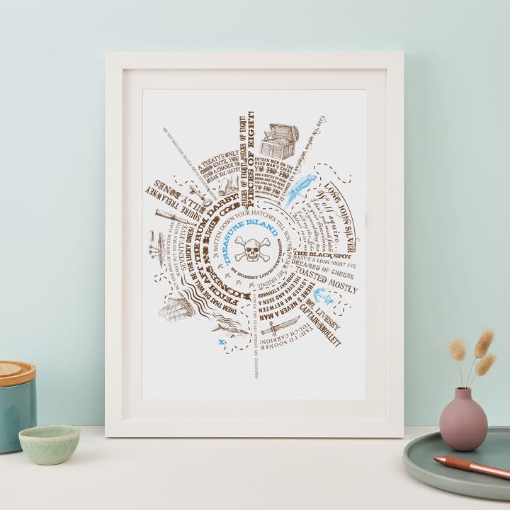 A framed print with a circular design featuring pirate quotes and illustrations is displayed on a table. Next to it are a small plant pot, a bowl, a vase with dried flowers, and a pen.