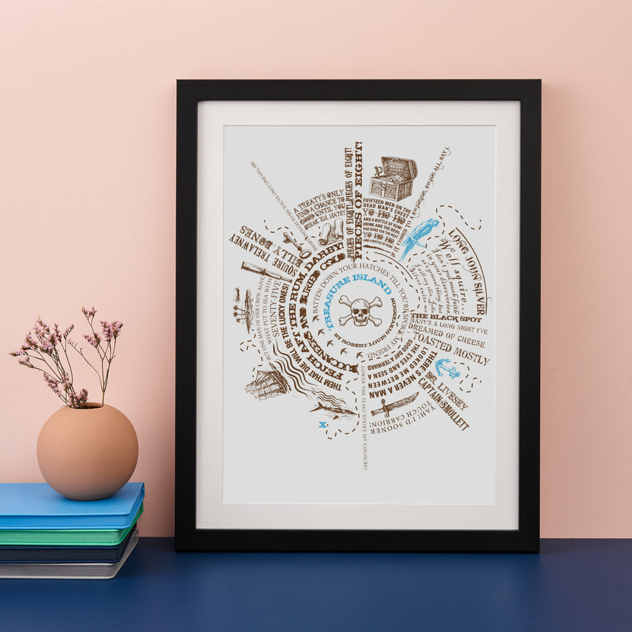 A framed print featuring a circular design with pirate-themed quotes and illustrations is displayed on a table. A small vase with a sprig of dried flowers and a stack of books sit next to the frame.