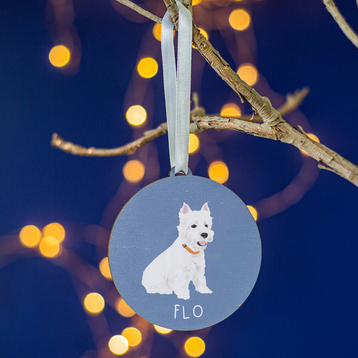 A round wooden ornament featuring a detailed hand-painted image of 'Flo,' a white dog, set against a cozy backdrop of glowing Christmas lights.