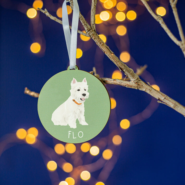A personalized green color Christmas ornament with a hand-painted portrait of a white dog named 'Flo,' elegantly hanging from a branch amid a twinkling holiday ambiance.