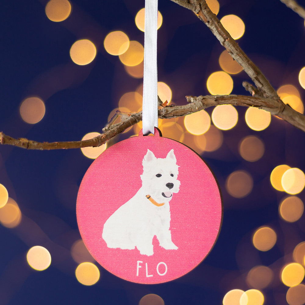 A personalized Pink Christmas ornament with a hand-painted portrait of a white dog named 'Flo,' elegantly hanging from a branch amid a twinkling holiday ambiance.