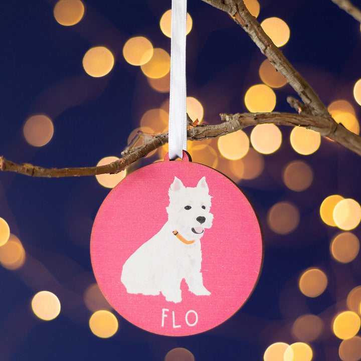 A personalized Pink Christmas ornament with a hand-painted portrait of a white dog named 'Flo,' elegantly hanging from a branch amid a twinkling holiday ambiance.