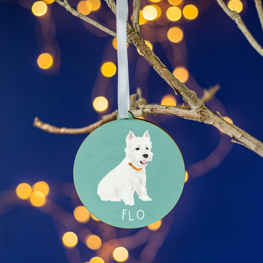 A round,teal coloured wooden ornament featuring a detailed hand-painted image of 'Flo,' a white dog, set against a cozy backdrop of glowing Christmas lights.