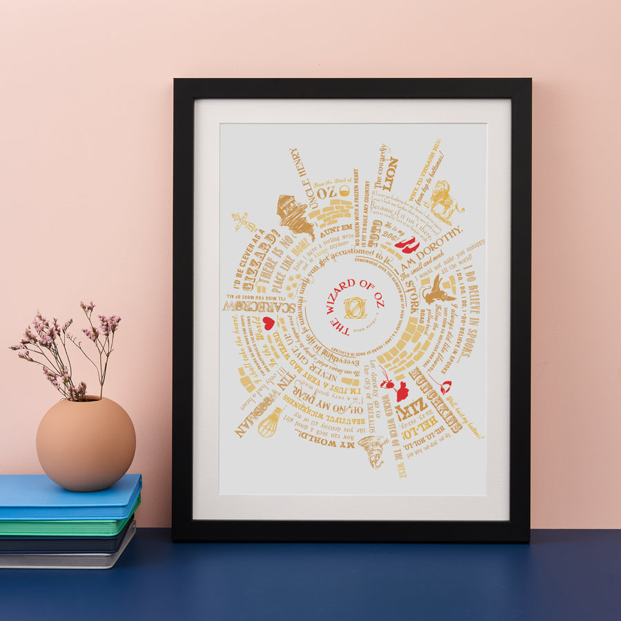 Framed typography art print of The Wizard of Oz in gold and red, displayed on a dark blue surface against a soft pink wall, with a small vase and stacked notebooks nearby.