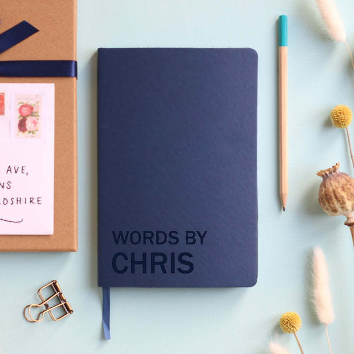 A top down image of a navy blue vegan leather resting on a pale aqua table. The front of the notebook is  engraved with the phrase Words By and personalised with the name Chris. Surrounding the book are various stationary items and dried flowers.