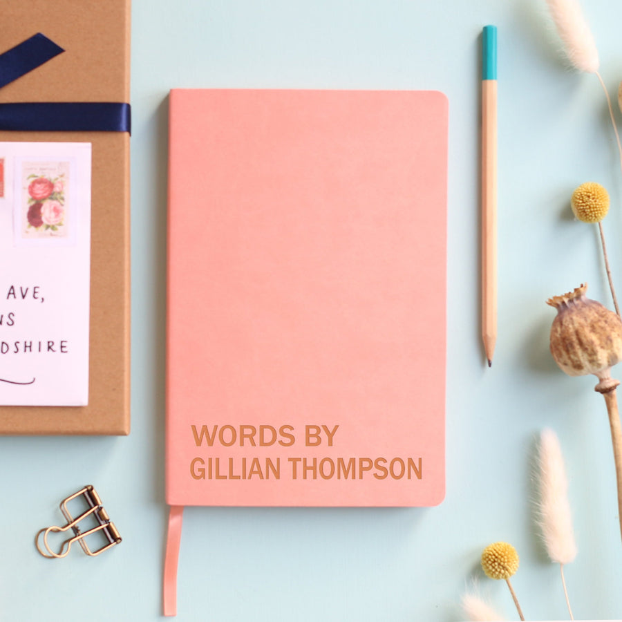 A top down image of a pink vegan leather resting on a pale aqua table. The front of the notebook is  engraved with the phrase Words By and personalised with the name Gillian Thompson. Surrounding the book are various stationary items and dried flowers.