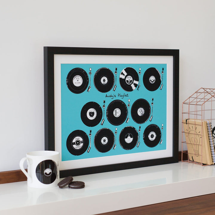 A framed print featuring a collection of vintage vinyl records playing on record players. The title "Andy's Playlist" is displayed at the bottom. A mug with a record design sits on a table nearby.
