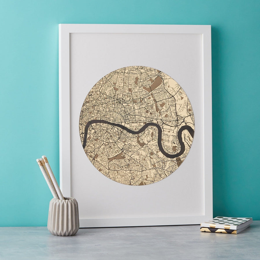A framed circular map of London with the Thames River winding through it, displayed in sepia tones. Pencils in a gray holder and a small book are on the table next to the frame.