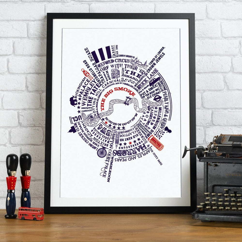 A framed print with a circular map of London landmarks and phrases, including "OXFORD CIRCUS," "THE SHARD," and "PUT A SMILE ON YER BOAT ME OLE CHINA." The print is in a black frame and sits on a wooden surface with toy soldiers, a toy bus, and a typewriter.