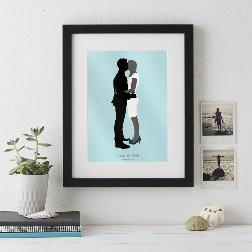  A wedding silhouette portrait in a black frame. The bride and groom are kissing on a light blue background. Their names and wedding date are written below.