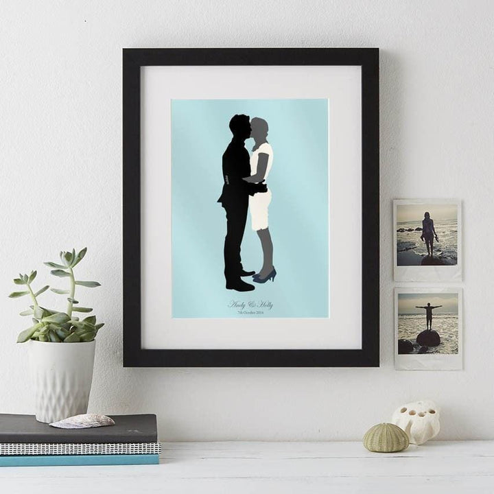  A wedding silhouette portrait in a black frame. The bride and groom are kissing on a light blue background. Their names and wedding date are written below.
