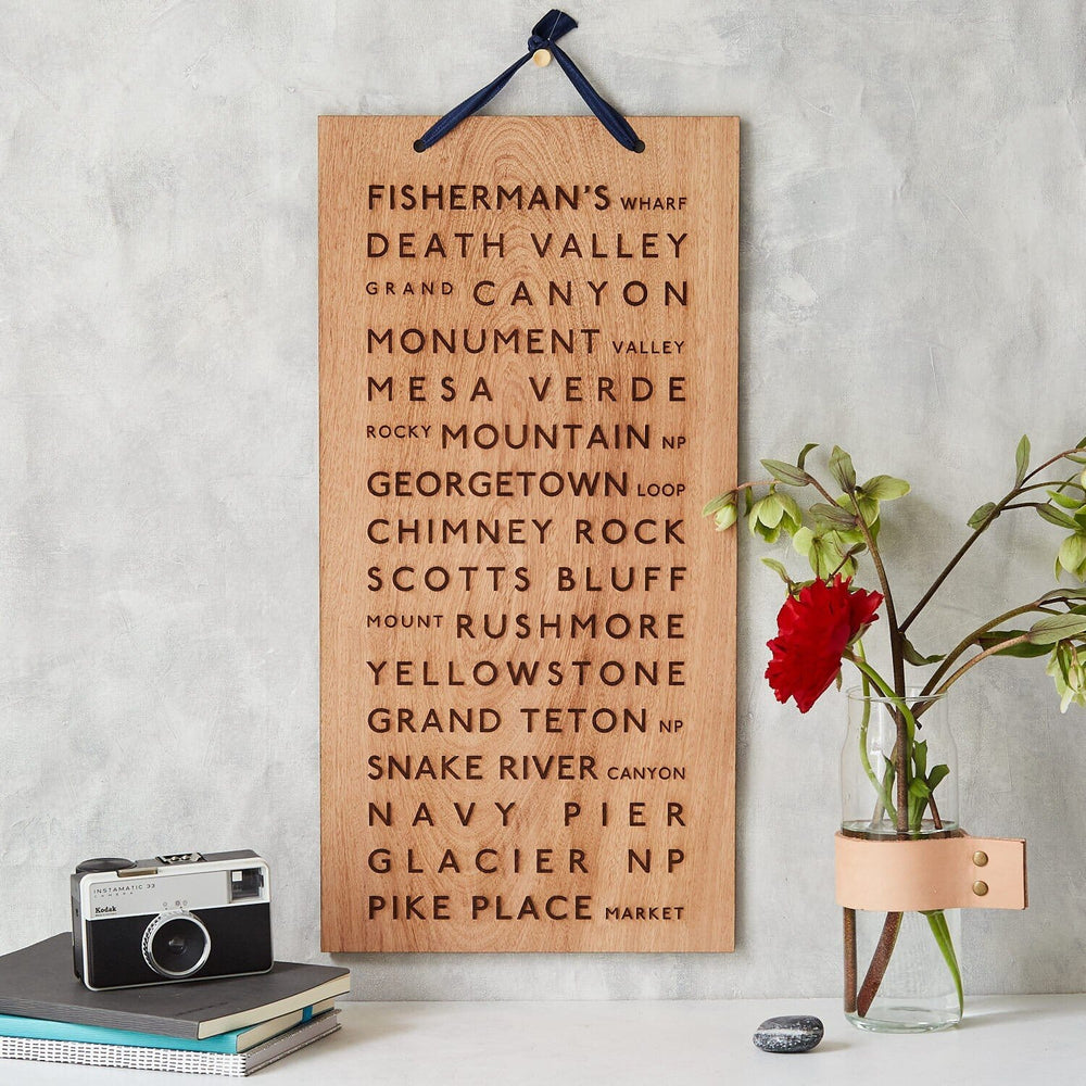 A wooden plaque featuring engraved names of iconic travel destinations, including the Grand Canyon and Yellowstone National Park, displayed alongside a camera, books, and a vase of flowers, creating a travel-inspired vignette.