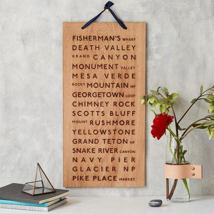 A wooden plaque featuring engraved names of iconic travel destinations, including the Grand Canyon and Yellowstone National Park, displayed alongside a vintage camera, a stack of books, and a vase of flowers, creating a cozy and inspiring atmosphere.