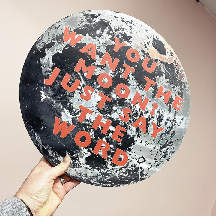 A round metallic moon-themed plaque with the quote "YOU WANT THE MOON? JUST SAY THE WORD" in bold orange letters, held by a hand against a neutral-colored background.