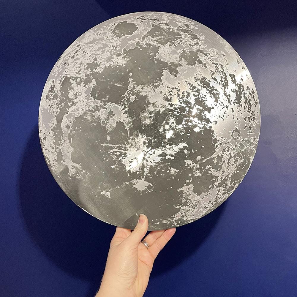 A hand holding a metallic moon-themed wall plaque against a deep blue background. The plaque reflects light, creating a glowing effect.
