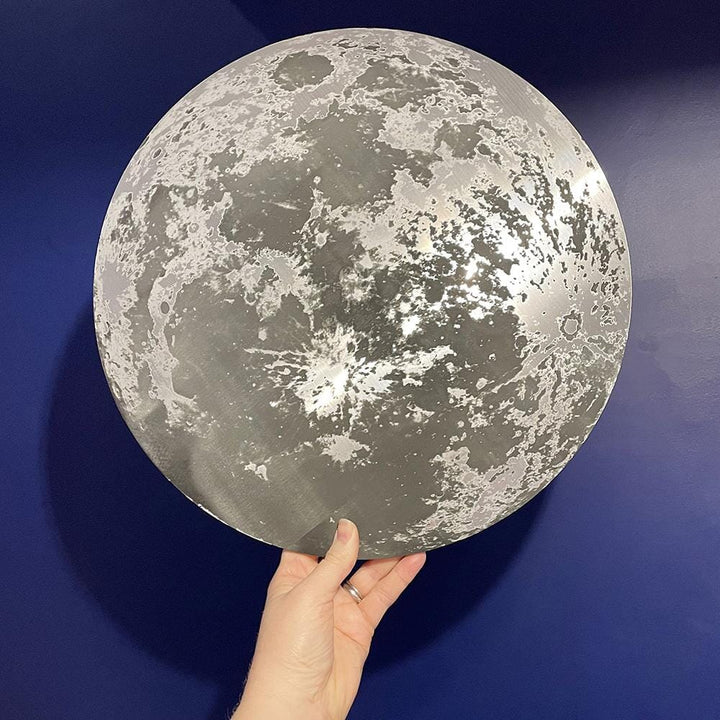 A hand holding a metallic moon-themed wall plaque against a deep blue background. The plaque reflects light, creating a glowing effect.