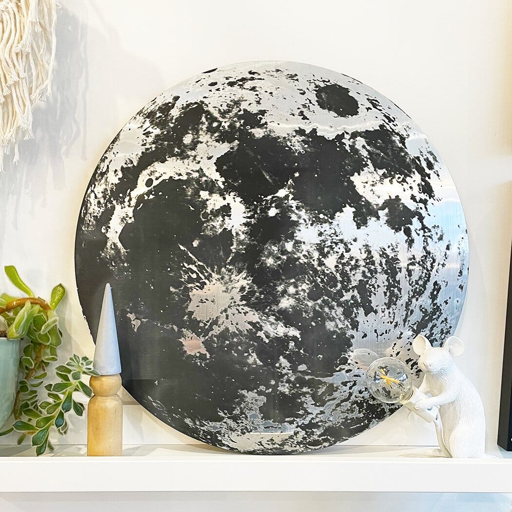 A metallic moon-themed wall plaque displayed on a shelf, surrounded by decorative items, including a small white mouse figurine holding a light bulb.
