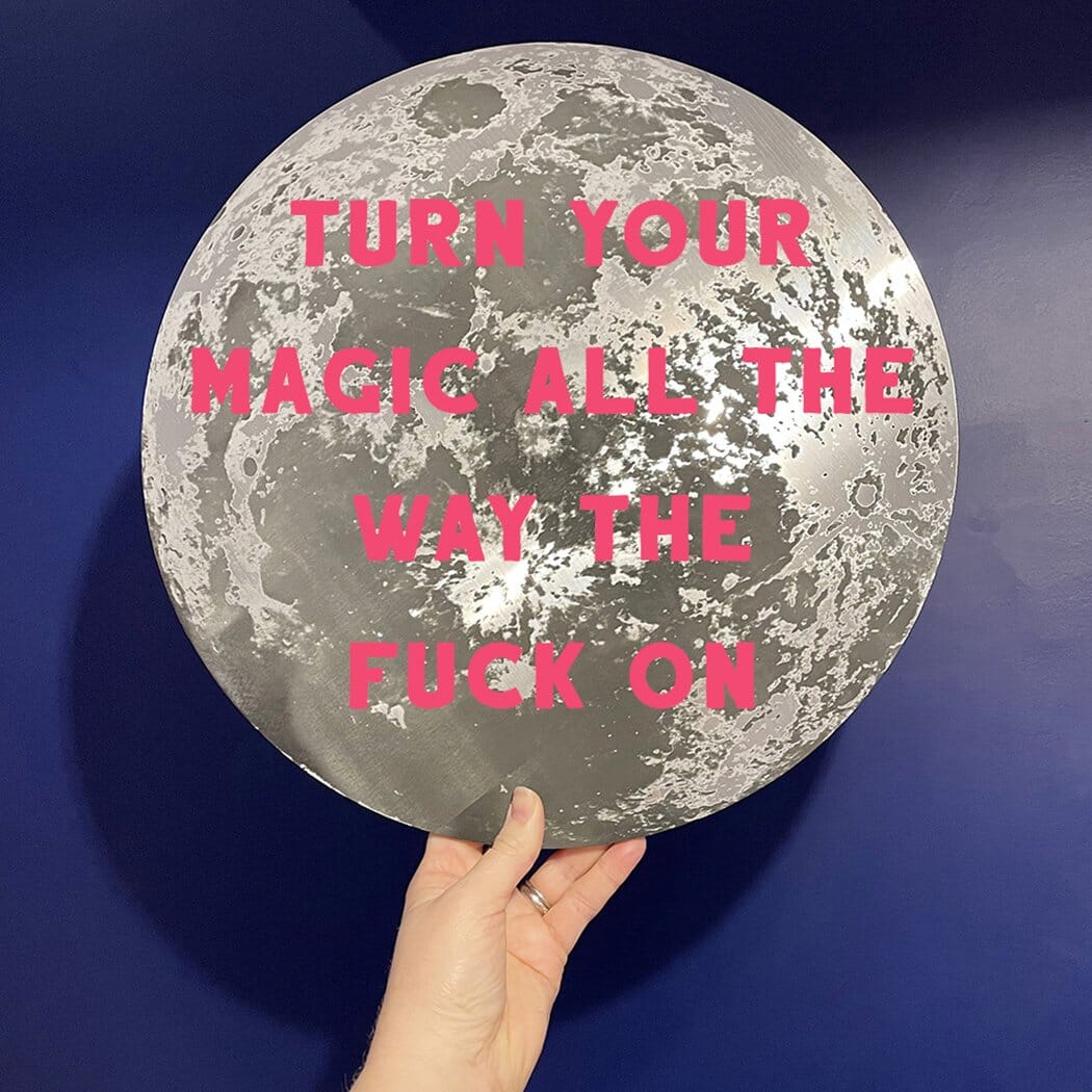 A round metallic moon-themed plaque with the quote "TURN YOUR MAGIC ALL THE WAY THE F*** ON" in bold pink letters, held by a hand against a dark blue wall.