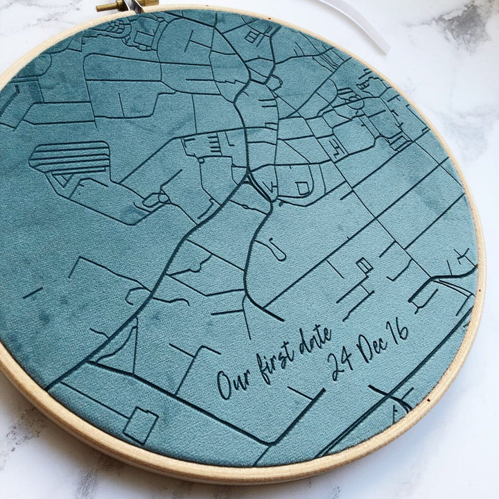 A close-up of an embroidered map in a circular hoop. The map is teal blue and features streets, rivers, and the text "Our first date 24 Dec 16."
