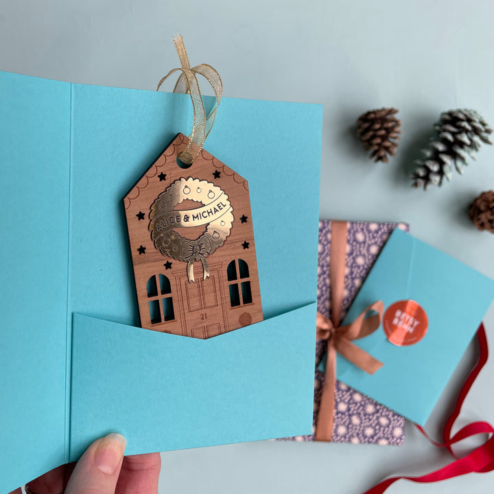 Wooden house Christmas ornament with personalised golden wreath and engraved details in a light blue gift sleeve. Makes a great secret santa gift.