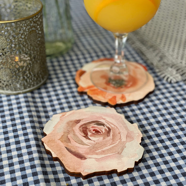 A close-up of a beautifully crafted wooden coaster featuring a hand-painted watercolor rose. The coaster is displayed on a checkered tablecloth, adding a touch of rustic charm.

