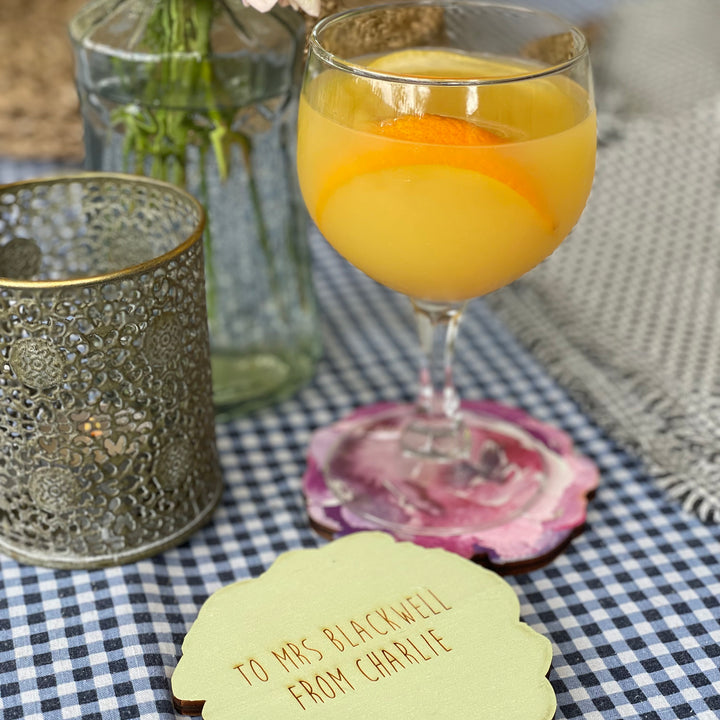 A wooden coaster with a personalized message "To Mrs. Blackwell from Charlie" displayed beside a glass of a refreshing cocktail.