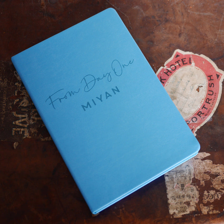 A top down image of a blue vegan leather notebook resting on a distressed tan leather suitcase. The front of the notebook features the phrase "From Day One". It is personalised with the name Miyan. 