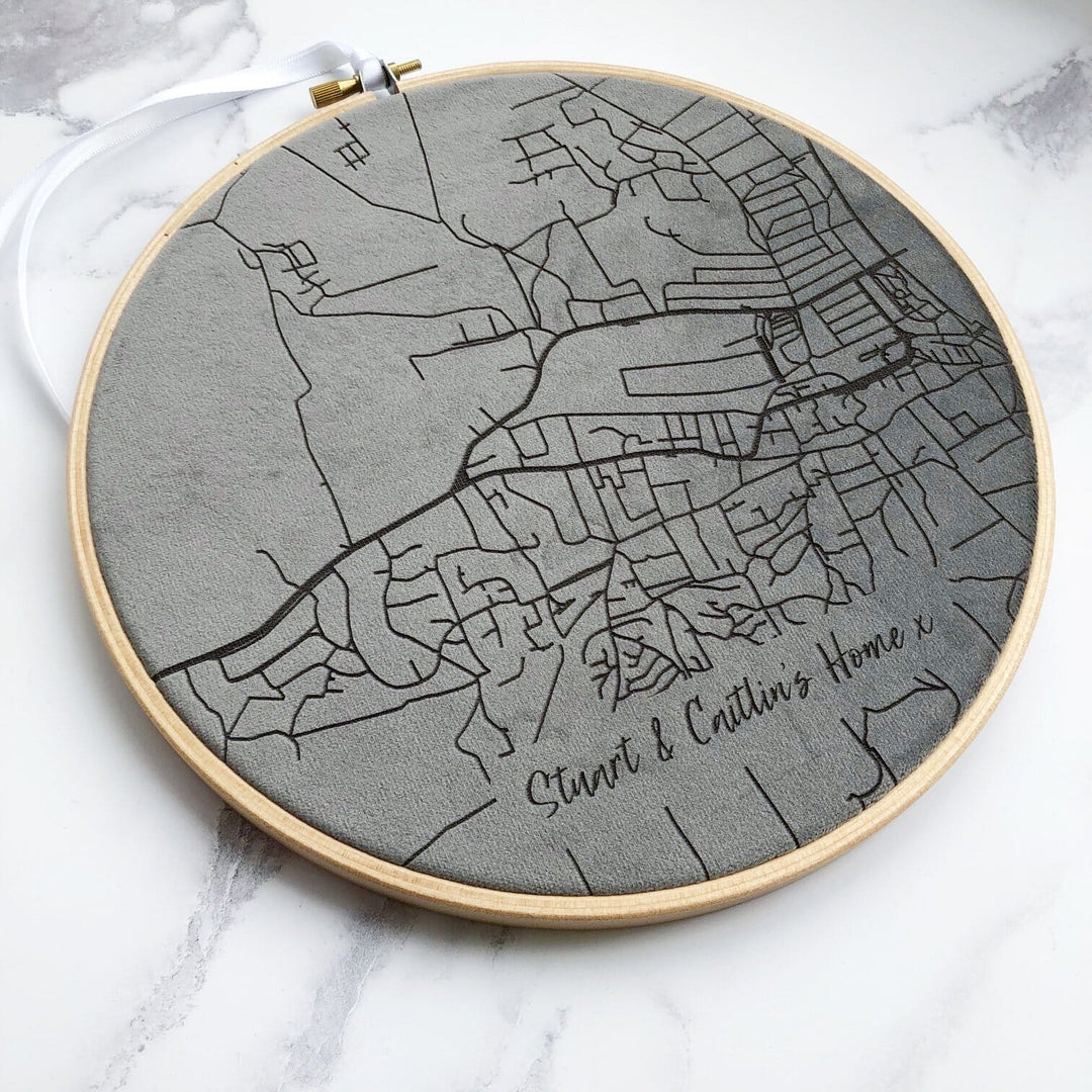 A circular embroidery hoop with a gray map of a city embroidered on it. The text "Stuart & Caitlin's Home" is embroidered at the bottom.