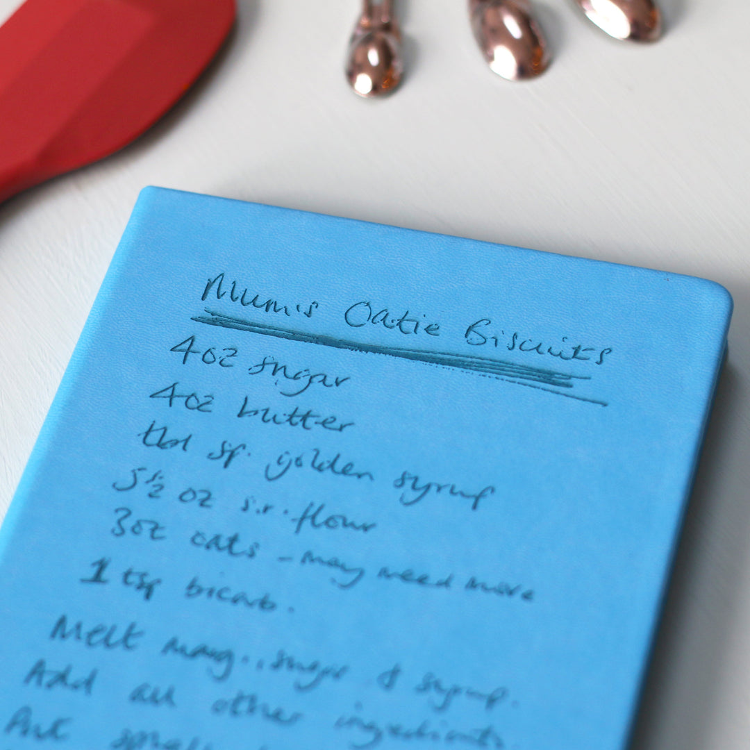 A close up image of a blue hardcover vegan leather notebook resting on a pale aqua table. The book is engraved with a handwritten recipe.