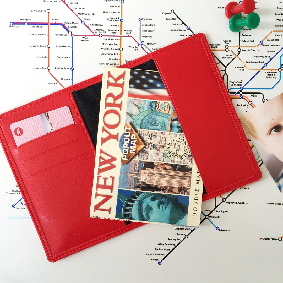 A red passport holder with a New York City pop-out map tucked inside, resting on a London Underground map. Two red and green paper pins are in the top right corner.