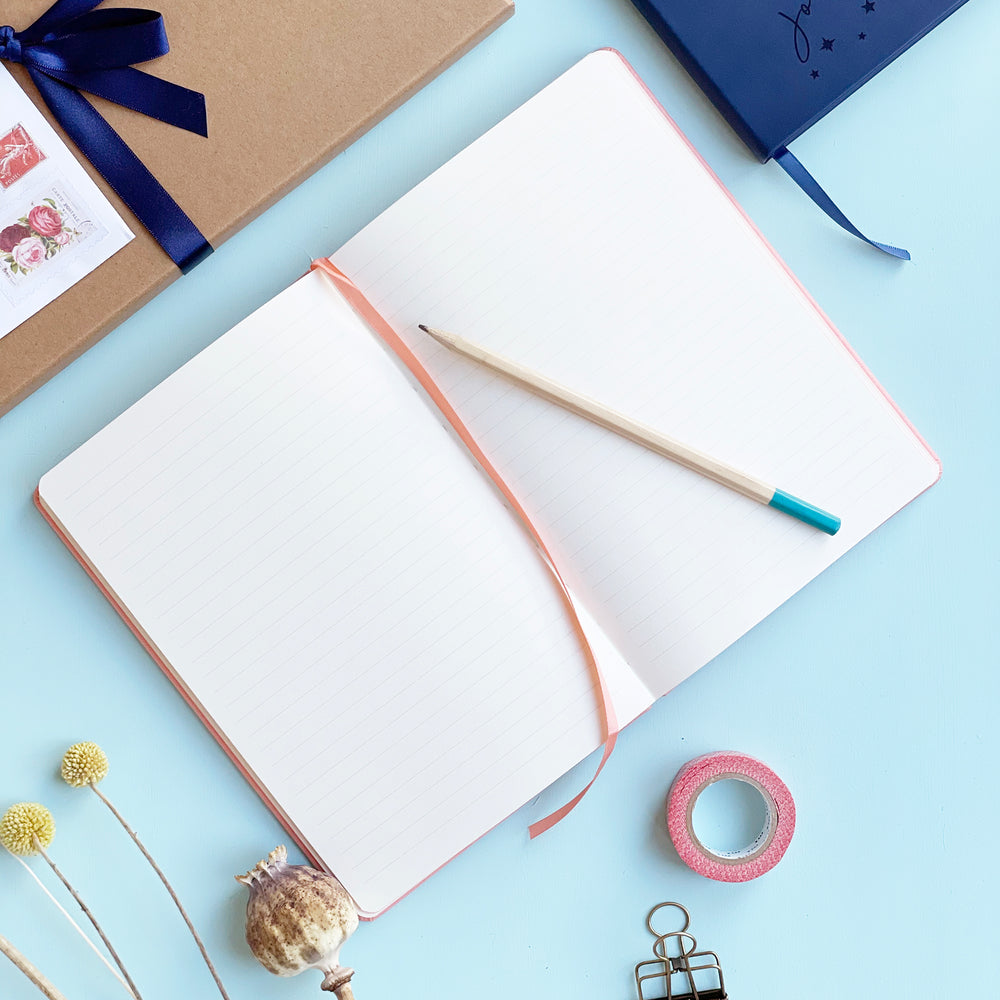 Personalised journal notebook lying flat open on pale aqua table to show lined pages and a matching colour ribbon place marker. A pencil is resting on the top of the notebook. Washi tape, clips and other stationery items are scattered around it. 