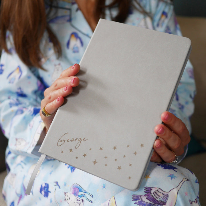 A close up image of a woman holding a grey vegan leather notebook. The front of the notebook is personalised with the name George above a cluster of different sized stars. 